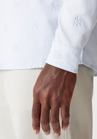 Camicia azzurro chiaro con loghi NY ricamati, dotata di polsini abbottonati con un accento logo. Il materiale sembra liscio e leggermente strutturato.