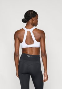 Vitt sport-bh med racerback-design, utrustad med en hake-och-hyska-stängning och ett utskuret detalj. Kombinerad med högmidjade svarta leggings.