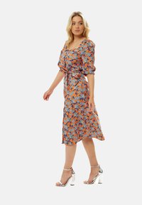 Floral dress with orange base, blue and white flower pattern, square neckline, puff sleeves, and knee-length hem paired with silver heeled sandals.