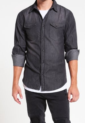 Mężczyzna w ciemnoszarej koszuli jeansowej zapinanej na guziki z podwiniętymi rękawami i czarnych spodniach, biała podkoszulka widoczna przy krawędzi i kołnierzu.