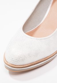 Gros plan sur une chaussure plate blanche texturée avec un embout arrondi et une semelle superposée marron et grise sur fond blanc.