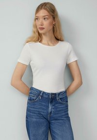 Witte geribde top met korte mouwen en een ronde halslijn, gecombineerd met vervagende blauwe high-waisted jeans. De stof lijkt licht en textuur te hebben.