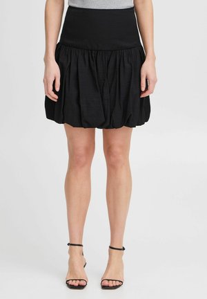 Gonna nera a palloncino con vita aderente, tessuto testurizzato e design arricciato. Abbinata a sandali neri con lacci, che mettono in mostra le gambe scoperte.