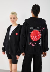 To personer iført svarte hettegensere med rosa kirsebærblomster og rødt sol-design; den ene står vendt mot kameraet, mens den andre viser detaljerte trykk på ryggen.