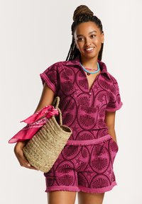 Krátký rukáv růžová vzorovaná polo košile s odpovídajícími šortkami, zdobená třásněmi, v kombinaci s tkanou kabelkou a růžovou šálou.