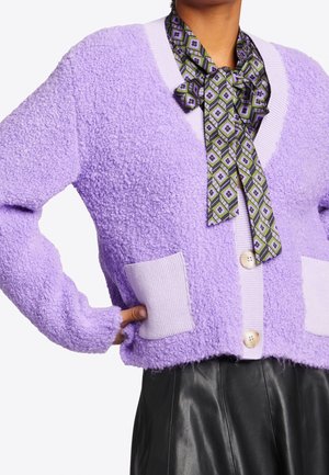 Persoon draagt een lavendelkleurige cardigan met textuur, twee voorzakken, grote knopen, een groen patroon hals sjaal en een zwarte leren broek.