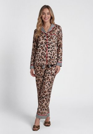 Vrouw staat, draagt satijnen pyjama met luipaardprint en zwart-wit gestreepte manchetten en kraag, glimlacht met handen ontspannen langs haar zij.
