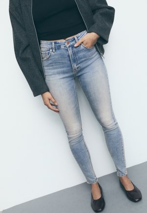 Osoba nosící vybledlé úzké modré džíny, černý top, šedou bundu a černé baleríny, stojící s jednou rukou v kapse opřená o bílou zeď.