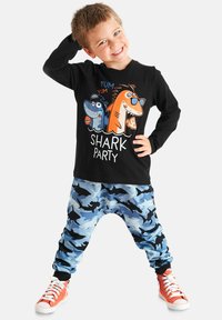 Sort langærmet skjorte med tegneseriehaj grafik; blå camo-stil bukser med haj silhuetter; orange sneakers med hvide snørebånd.