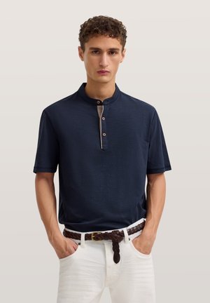 Junger Mann, der ein marineblaues Henley-T-Shirt mit Knöpfen und kurzen Ärmeln, weiße Hose und einen geflochtenen braunen Gürtel trägt, steht mit den Händen in den Taschen.