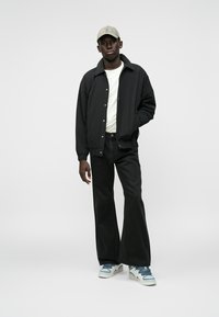 Jauns vīrietis, kurš valkā beigtu cepuri, melnu virsjak, baltu krekli, platās melnas bikses un baltus-zilus sporta apavus, stāv pret vienkāršu baltu fona.