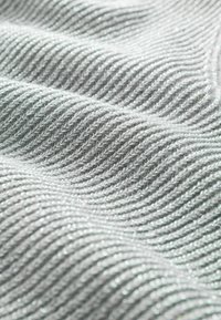 Hellgrauer gestrickter Stoff mit einem strukturierten, wellenförmigen Muster und subtiler Schimmerung, der gerippte Linien zeigt, die Tiefe und Dimension verleihen.