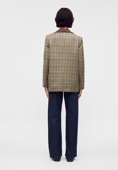 Personne aux cheveux courts et foncés, vue de dos, portant un blazer marron à carreaux et un pantalon large foncé, sur un fond clair uni.