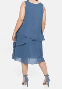 Robe en chiffon bleu à design superposé, sans manches, avec fermeture éclair au dos. Associée à des chaussures à talons hauts bleu clair, avec brides à la cheville.