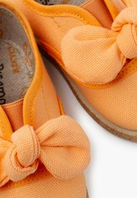 Scarpe in tela arancione con tomaia texturizzata, dotate di un grande nodo decorativo sulla punta e cuciture a contrasto lungo i bordi.