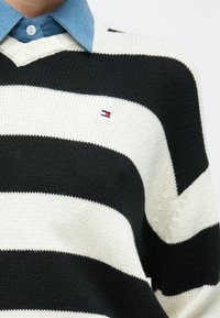 Πλεκτό πουλόβερ με μαύρες και λευκές ρίγες, με λαιμόκοψη σε σχήμα V και μικρό λογότυπο. Διαθέτει υφή υφάσματος και από κάτω φορεμένο πουκάμισο τζιν σε στρώσεις.