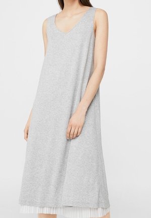 Vestito senza maniche grigio chiaro a scollo a V con vestibilità ampia, sovrapposto a una gonna bianca plissettata, indossato da una persona in piedi su uno sfondo semplice.