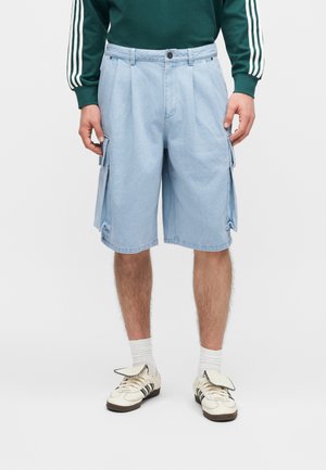 Homme portant un short cargo bleu clair, des baskets blanches avec des rayures noires, des chaussettes blanches et un haut vert à manches longues avec des rayures blanches sur les manches.