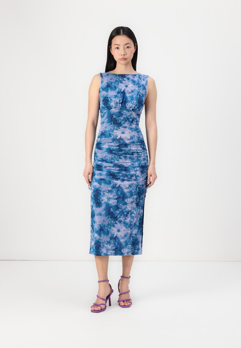 Robe florale bleue cintrée avec des détails froncés, sans manches, longueur genou, associée à des talons strappy violets. Texture du tissu lisse.