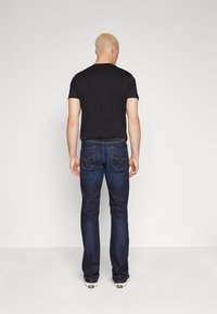 Άντρας με κοντά ξανθά μαλλιά στέκεται μπροστά σε έναν λευκό τοίχο, φορώντας μαύρη μπλούζα, σκούρο μπλε τζιν και λευκά αθλητικά παπούτσια.