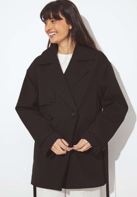 Cappotto nero doppiopetto con colletto ampio, maniche oversize e grandi bottoni. Tessuto liscio e texturizzato. Legaccio regolabile in vita.