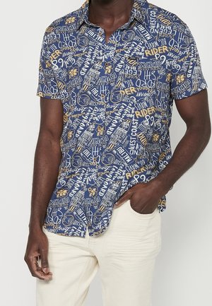 Homme portant une chemise bleue à manches courtes boutonnée avec un texte beige et des motifs floraux, associée à un pantalon blanc cassé.