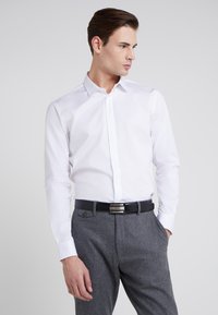 Camisa blanca de manga larga con cuello puntiagudo, combinada con pantalones grises texturizados y un cinturón negro con hebilla de plata.