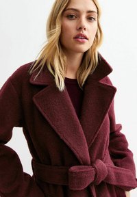 Cappotto in misto lana bordeaux con ampio colletto a punta, cintura a fiocco e texture morbida. Presenta maniche oversize e una vestibilità rilassata.
