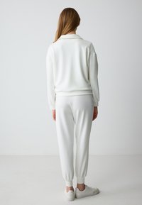 Witte lounge set bestaande uit een langermige, met kraag, top en elastische broek met cuffs. Zachte textuur, minimalistisch ontwerp, gecombineerd met witte sneakers.