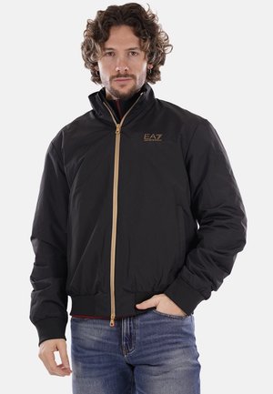 Black bomber jacket with a zippered front, gold accents, and ribbed cuffs. Features a high collar and elastic waistband for a fitted look.