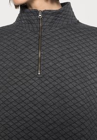 Donkergrijze rits-hals trui gemaakt van textuurstof met een ruitpatroon. Beschikt over een hoge kraag en metallic ritsdetail.