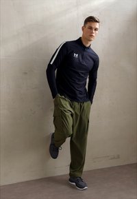 Fekete, hosszú ujjú, cipzáras ing fehér vállcsíkokkal, olíva zöld cargo nadrág és fekete sportcipő, semleges háttér előtt.