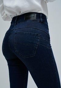 Närbild av en person som bär mörkblå slim-fit jeans med bakfickor och en svart läder-logotyp på midjebandet.