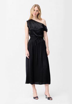 Mujer con top de satén negro de un solo hombro, falda midi negra con cintura ajustable y sandalias negras de tacón con puntera abierta, de pie sobre fondo blanco.