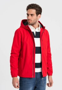 Giacca rossa con cappuccio realizzata in tessuto liscio, dotata di zip frontale, tasche laterali e polsini elasticizzati. Indossata sopra una polo a righe.