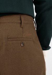Bruine wollen broek met een achterzak met een knoop en geborduurde merknaam. Glad textuur, op maat gemaakte pasvorm en klassiek ontwerp.