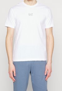 Mann in einem weißen T-Shirt mit kurzen Ärmeln und einem kleinen EA7 Emporio Armani-Logo auf der Brust, dazu blaue Hosen.