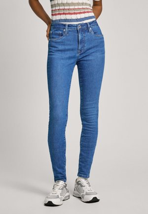 Jean skinny bleu en denim, comportant cinq poches, une fermeture à bouton et zip, et un délavage légèrement usé. Porté avec des baskets sportives.