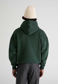 Persona vistiendo una sudadera con capucha de color verde oscuro, un gorro de punto color crema, y pantalones de rayas grises, de pie de espaldas contra un fondo liso.