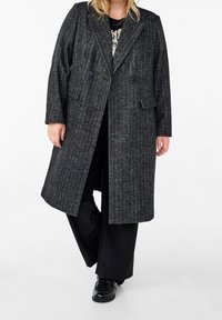 Dvojřadový šedý kabát z herringbone vzoru s klopovým límcem, bočními kapsami a texturovanou tkaninou. Nosený přes černý outfit a boty.