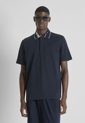 Jeune homme portant des lunettes de soleil foncées et un polo bleu marine à manches courtes avec un col à motif, debout devant un fond uni.