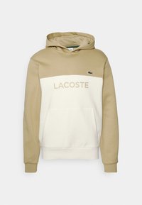 Sudadera con capucha en beige y crema, con bolsillo tipo canguro, logo "LACOSTE" en beige y pequeño emblema de cocodrilo verde en el lado izquierdo del pecho.