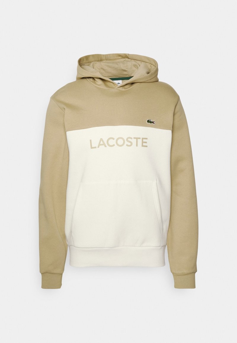 Sudadera con capucha en beige y crema, con bolsillo tipo canguro, logo "LACOSTE" en beige y pequeño emblema de cocodrilo verde en el lado izquierdo del pecho.