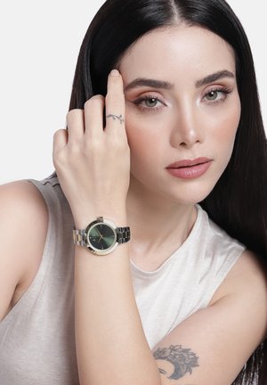 Pikkade mustade juustega naine kannab ilma varrukateta beežitoppi, millel on käes hõbedane käekell tumesinise näo ja nähtavate tätoveeringutega käel ja käel.