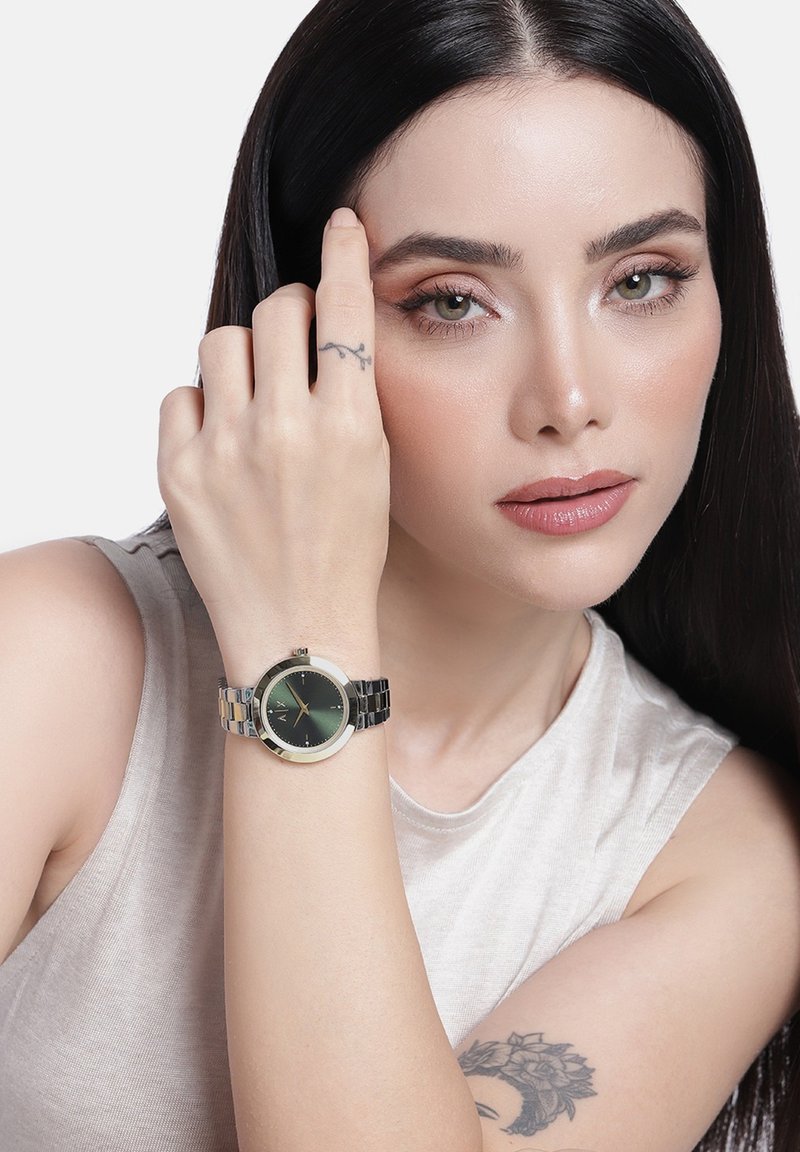 Pikkade mustade juustega naine kannab ilma varrukateta beežitoppi, millel on käes hõbedane käekell tumesinise näo ja nähtavate tätoveeringutega käel ja käel.