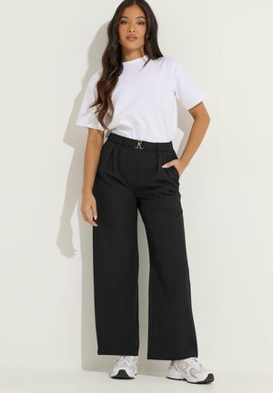 Kobieta z długimi włosami ubrana w białą koszulkę, czarne, szerokie spodnie z wysokim stanem, białe trampki, pozuje z jedną ręką w kieszeni.