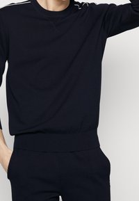 Homme portant un sweat-shirt à manches longues noir et un pantalon assorti, une main dans la poche, sur un fond clair uni.