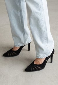 Persona que lleva vaqueros azul claro y tacones altos negros con punta puntiaguda y detalle de malla, de pie sobre un suelo gris liso.