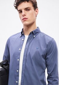 Modrá bavlnená košeľa na gombíky s klasickým golierom, s malým logom na hrudi a kontrastnými bielymi gombíkmi. Doplnkom je čierna taška.
