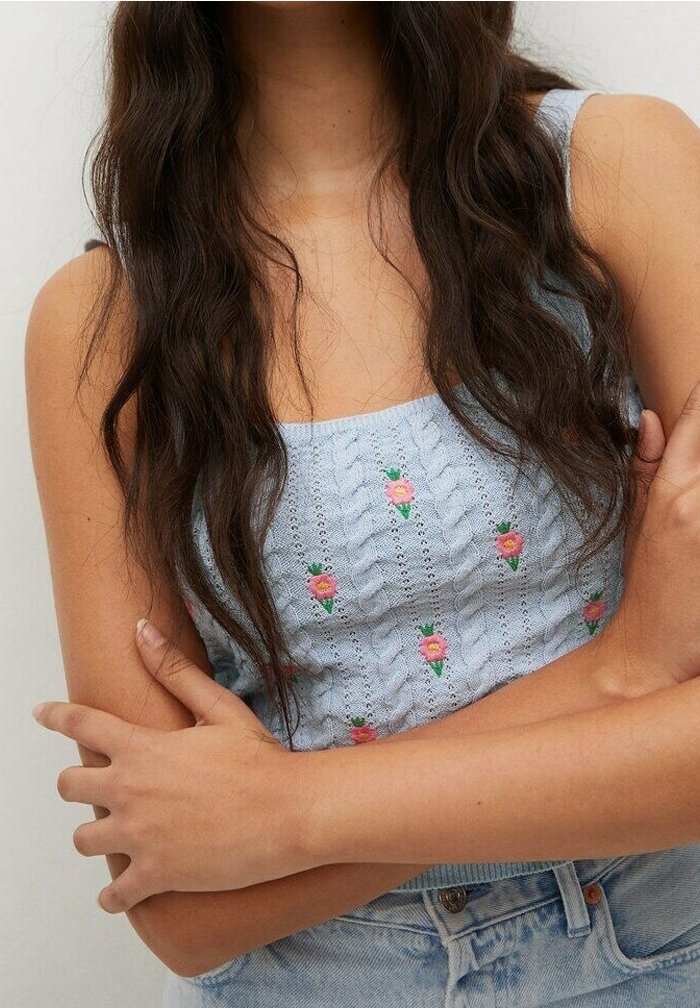 Femme aux longs cheveux foncés porte un débardeur en maille bleu clair avec de petits motifs floraux roses et un jean en denim bleu clair, les bras croisés.